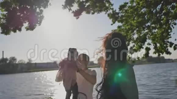 一个三口之家在阳光照射下的湖岸绿树下爸爸高高举起他的小手视频的预览图