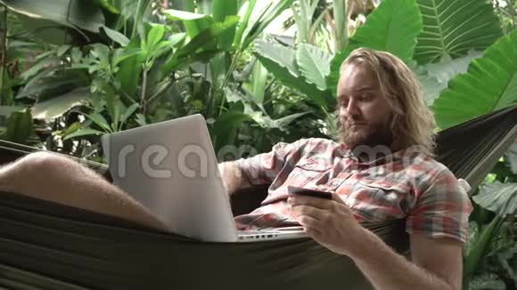 男人坐在吊床上成功地用信用卡和笔记本电脑付款视频的预览图