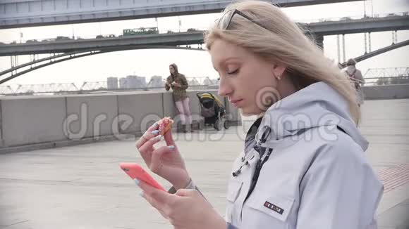 一位年轻漂亮的金发女郎在河堤的长凳上放松时吃了一个甜甜圈享受着她的智能手机视频的预览图