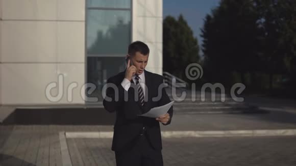 穿着黑色西装的商人在智能手机上交谈并在商业建筑背景下向户外扔文件视频的预览图