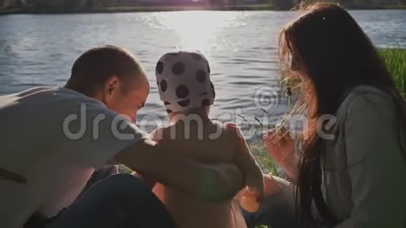 一个年轻的家庭三个人在湖岸的夕阳下公园里阳光明媚的夜晚父母视频的预览图