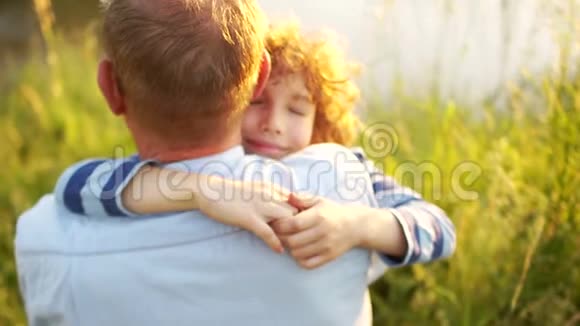 男孩和他成熟的父亲坐在河岸上日落儿子紧紧地搂着父亲的肩膀视频的预览图