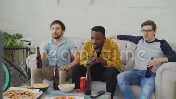 一群年轻的男性朋友一起在电视上看体育比赛一边在家喝啤酒一边吃零食视频的预览图