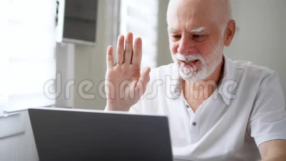 老人坐在家里拿着笔记本电脑使用电脑聊天通过信使应用程序微笑着挥手致意视频的预览图