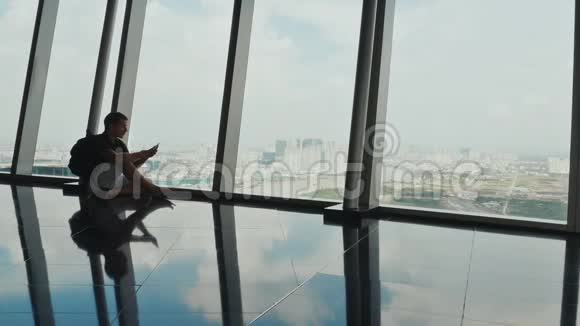 一位坐在摩天大楼窗户旁的游客的剪影并与电话一起工作视频的预览图