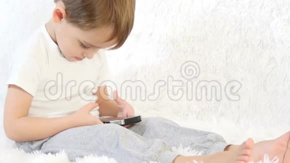 快乐的孩子在白色背景下玩智能手机一个孩子坐在白色沙发上特写镜头视频的预览图