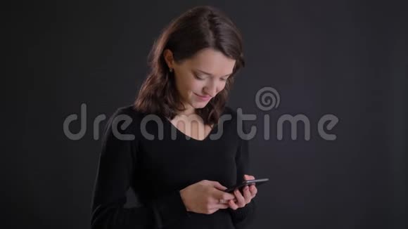 年轻迷人的白种人长发女孩在黑色背景下微笑地观看智能手机的肖像视频的预览图