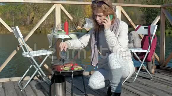 年轻女子穿着暖和的衣服在烤架上准备蔬菜和肉用电话在河岸上野餐视频的预览图