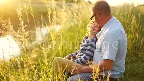 祖父和孙子坐在河岸上成熟的男人和卷发的男孩在草地上放松视频的预览图