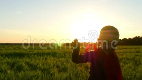 日落时一个穿着超级英雄服装穿着红色斗篷的快乐孩子穿过一片绿色的草坪视频的预览图