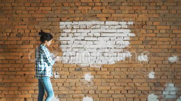 一个年轻的女孩用滚筒在她的房子里画一堵砖墙用滚筒粉刷墙壁把裸露的墙壁漆成视频的预览图