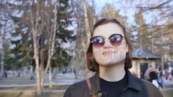 城市公园里戴太阳镜微笑的白种人年轻女子的肖像慢动作视频的预览图