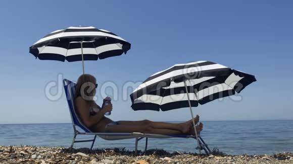 穿比基尼的美丽成年女人躺在海滨的躺椅上喝水视频的预览图