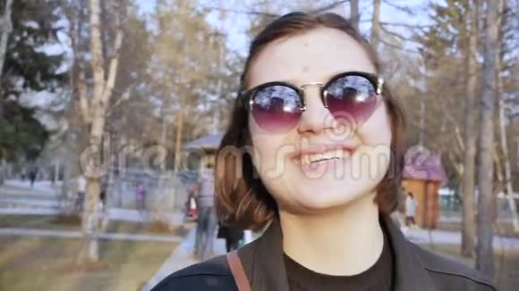 城市公园里戴太阳镜微笑的白种人年轻女子的肖像慢动作视频的预览图