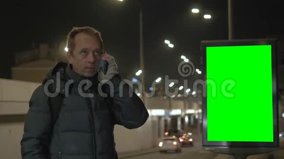 一个男人在城里晚上打电话在背景下有一个带有绿色屏幕的广告牌视频的预览图