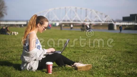 一个女孩坐在公园里草地上在视频交流中交谈一个学生一个自由职业者微笑和微笑每杯咖啡视频的预览图