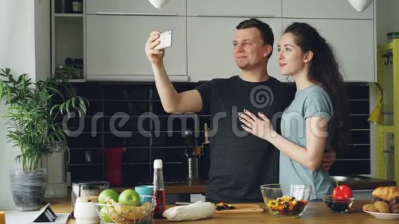 一对年轻有趣的夫妇早上在家厨房做饭时用智能手机相机拍自拍照片视频的预览图