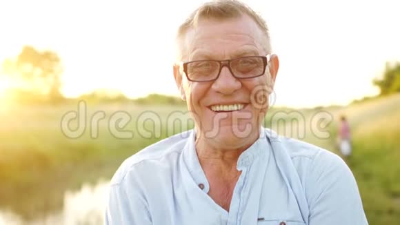 一位微笑开朗的退休人员的特写肖像一个戴着光学眼镜的人在夕阳田野和河流中视频的预览图