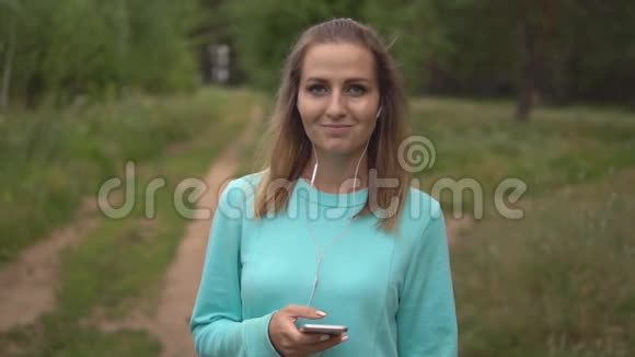 美丽的年轻女孩手里拿着手机带着耳机听音乐在室外相机微笑视频的预览图