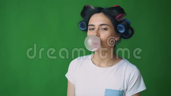 女孩头上戴着卷发器嚼着口香糖在绿色背景上吹泡泡发色视频的预览图