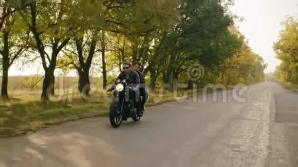 秋天在森林路上骑摩托车的年轻酷夫妇的追踪镜头戴太阳镜的漂亮小伙子视频的预览图