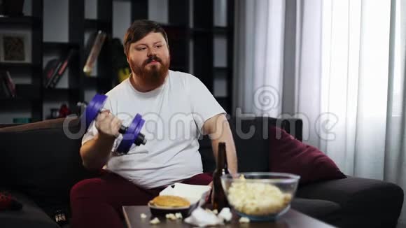 有趣的胖子吃快餐喝啤酒做哑铃训练他正坐在家里的沙发上视频的预览图