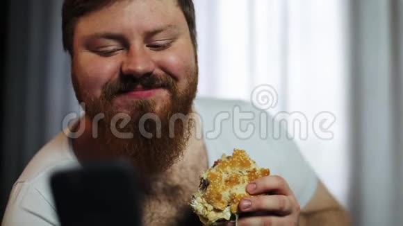 一个长着大胡子的胖子在他的智能手机里打东西在沙发上吃汉堡视频的预览图