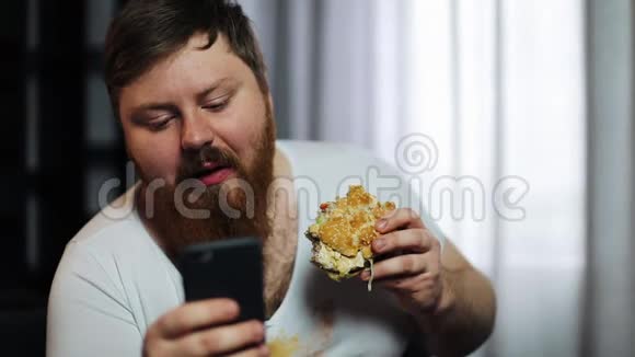 一个长着大胡子的胖子在他的智能手机里打东西在沙发上吃汉堡视频的预览图
