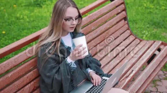 一个年轻的学生带着笔记本电脑在夏天的公园里坐在长凳上看书喝杯咖啡视频的预览图