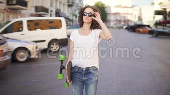 快乐的女人摸着她的太阳镜然后开始走路拿着她时尚的长板在早上慢镜头视频的预览图