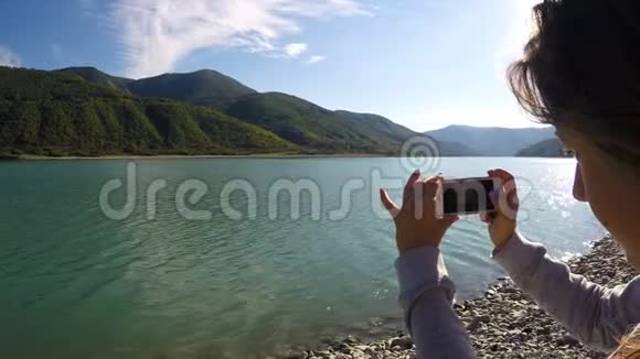 年轻女士用智能手机录制阳光山景和河流的视频视频的预览图