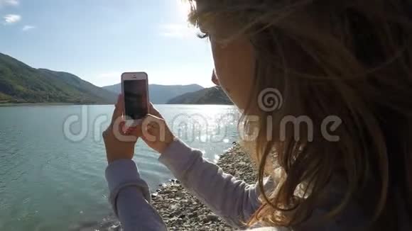 阳光明媚的一天女性用手机拍摄山景和河流视频的预览图