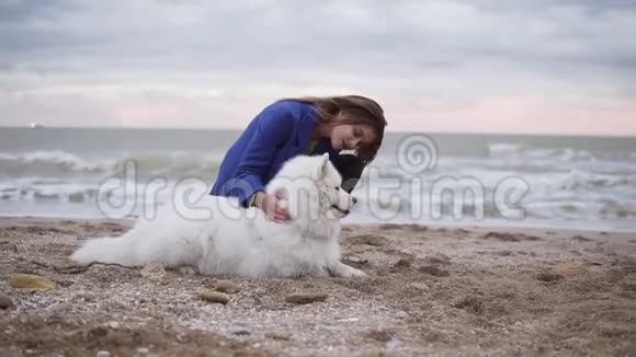 圆形镜头动作年轻的女人坐在沙滩上拥抱她在海边的萨莫伊德品种的狗白色视频的预览图