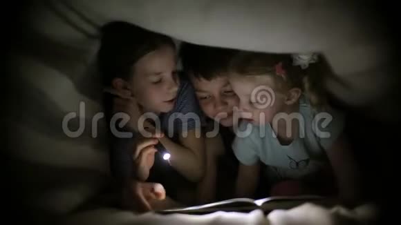 两个姐妹和一个兄弟在一个黑暗的房间里用手电筒在毯子下读一本书孩子们在玩视频的预览图