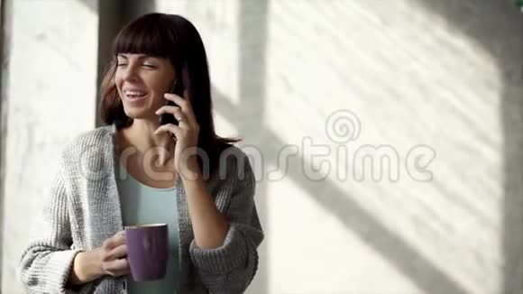 一位美丽的女士坐在窗户附近喝热茶她在电话里讲话视频的预览图