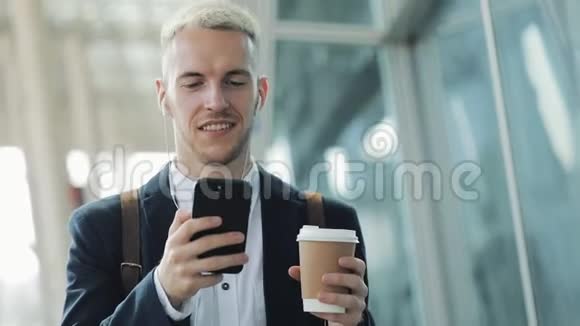 年轻有魅力的男人拿着咖啡和电话走在街上他用耳机听音乐走路视频的预览图