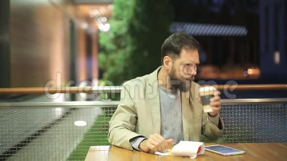 一个长着胡子的白种人中年男子晚上坐在夏季咖啡馆的一张桌子旁观看视频的预览图
