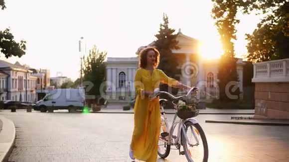 黎明时分一个身穿黄色长裙的微笑着的黑发女人手里拿着她的自行车带着鲜花的车把视频的预览图