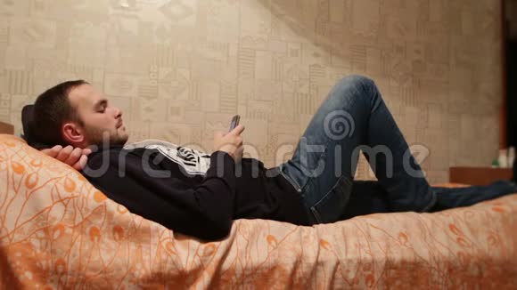 有胡子的懒汉放松地躺在家里的床上在肖像模式下使用手机懒惰的白种人视频的预览图