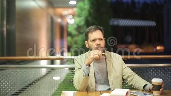 一个长着胡子的白种人中年男子晚上坐在夏季咖啡馆的一张桌子旁观看视频的预览图
