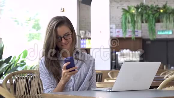 戴眼镜的女人用智能手机桌上的笔记本电脑视频的预览图