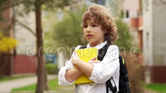 一个可爱的卷曲的小学生站在背后背着一个背包手里拿着一本书风吹着他的头发视频的预览图