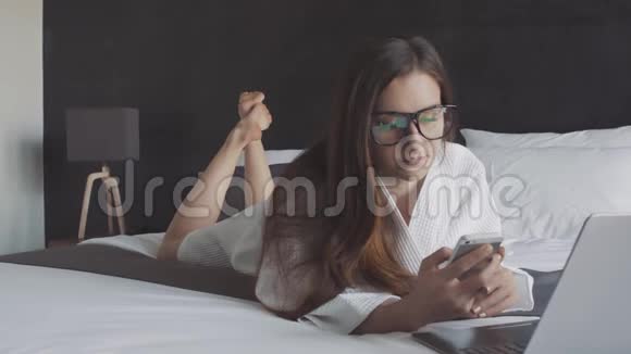 穿着白色浴袍戴着眼镜的女商人躺在床上用智能手机聊天用笔记本电脑工作在线商务视频的预览图