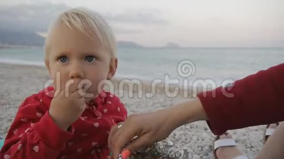 特写两岁的宝宝在海滩上吃一个铝箔托盘上的绿色橄榄孩子和妈妈在日落海滩上视频的预览图