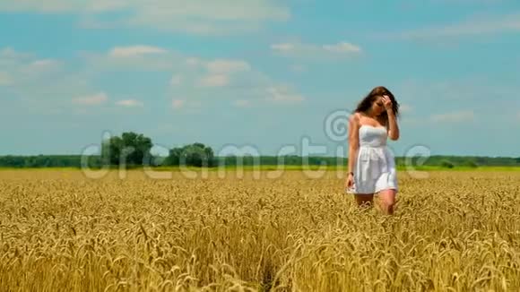 一位美丽的黑发年轻女子穿着白色短夏短裙在金色的麦田里散步视频的预览图