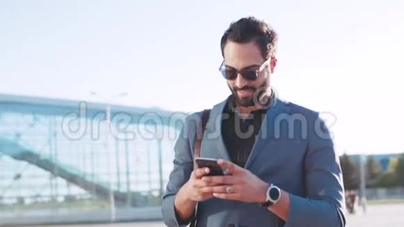 一位留着大胡子的时髦男士站在机场候机楼旁用手机旋转着迷人地微笑着视频的预览图