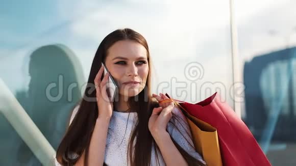 销售消费主义年轻女性手拿智能手机和购物袋站在购物中心附近聊天视频的预览图
