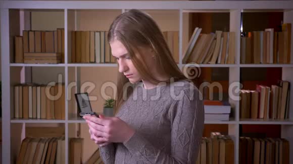 在室内的大学图书馆里用手机和照相机拍摄了一幅年轻迷人的女学生的特写肖像视频的预览图