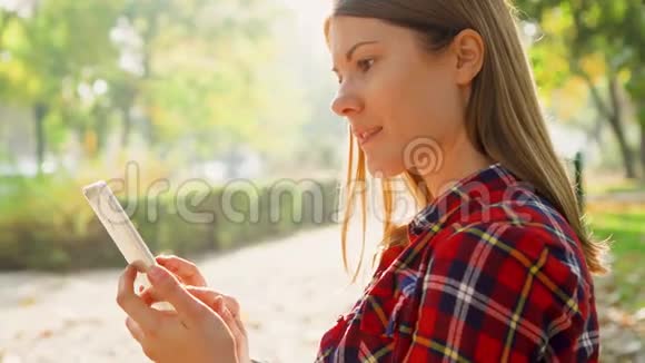微笑的女人享受大自然坐在绿色公园的长凳上通过信使应用程序用手机聊天视频的预览图