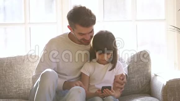 快乐的爸爸和小女儿坐在沙发上用智能手机视频的预览图